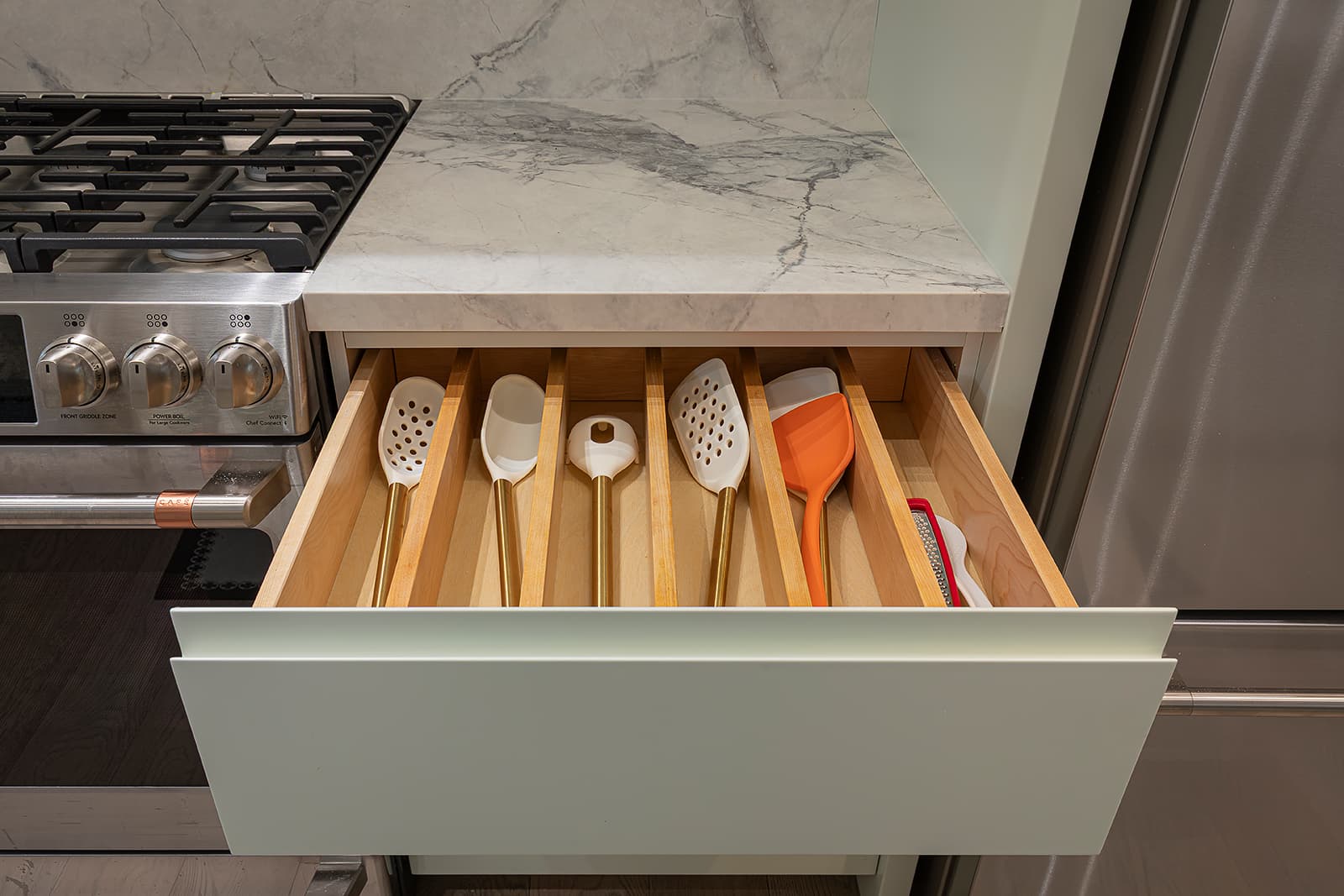 Open kitchen drawer with wooden utensil organizer holding spatulas and cooking tools beside a stainless steel stove and quartz countertop.