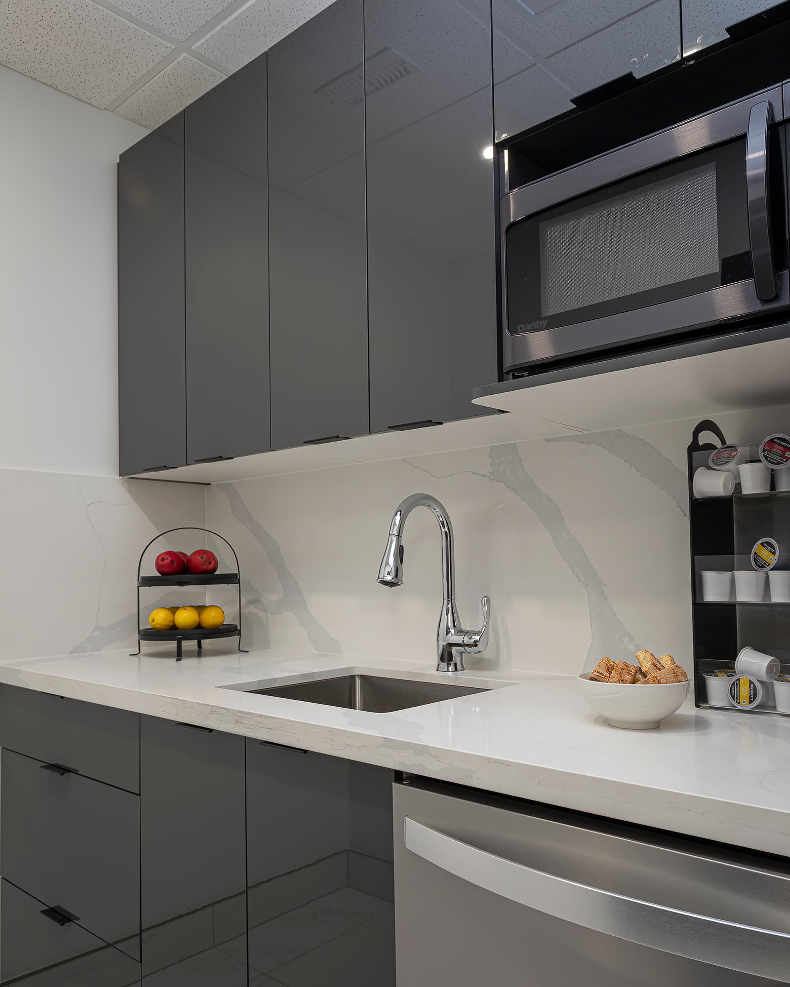 Modern kitchen with glossy grey cabinets, white quartz countertop, stainless steel sink, and built-in microwave with fruit and coffee accessories.